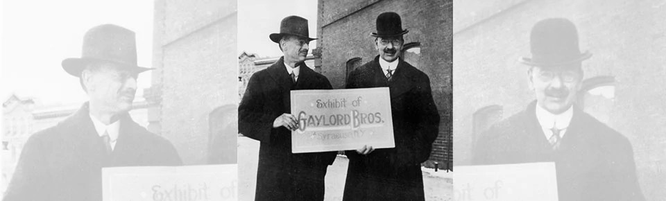 Black and white photo of two men wearing hats holding a sign "Exhibit of Gaylord Bros. Syracuse, NY"